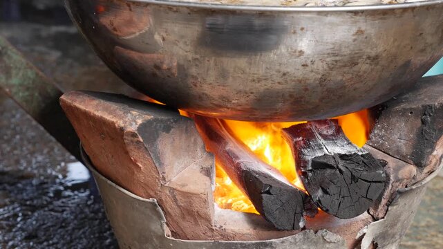 Large wok heating over open flame on a clay charcoal stove where fire and hot coals warm the metal surface in a traditional outdoor cooking setup. Street cuisine.