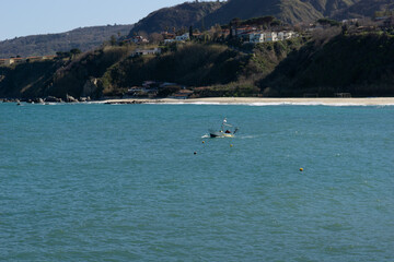 Obraz premium small fishing boat floating on calm turquoise mediterranean waters near the coastline of tropea italy, lush vegetation under a clear blue sky, concept of travel agency, coastal tourism, maritime life.