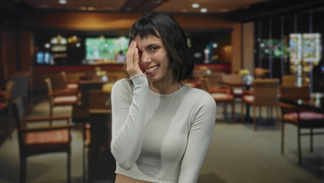 Young hispanic woman smiling broadly with hand covering face in a cozy restaurant building; embarrassment.