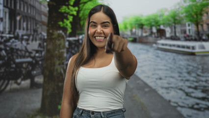 Fototapeta premium Woman pointing finger at camera on street canal path, smiling in white tanktop and jeans; joy confidence.