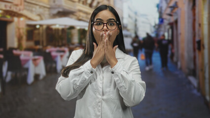Fototapeta premium Woman doctor in white coat covers mouth with hands in a surprised gesture on a street cafe scene; surprise empathy.
