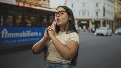 Fototapeta premium Young woman wearing glasses makes finger gun with hands aimed at camera on a busy city street with a red bus behind; playful confidence.
