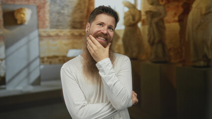 Obraz premium Bearded man with earrings touches his chin in a museum building among ancient marble statues under soft lighting; contemplation.