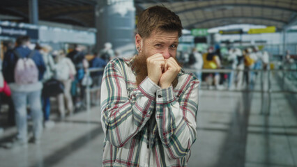 Fototapeta premium Young caucasian man covers mouth with fists while standing in crowded airport terminal check in queue; anxiety.