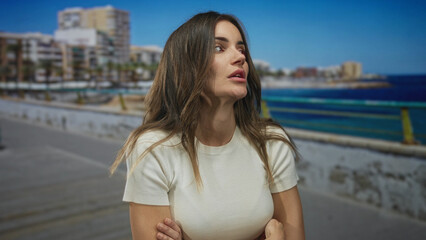 Fototapeta premium Young hispanic woman gazes sideways with finger to lips and bare arms crossed on seaside street; anxiety.