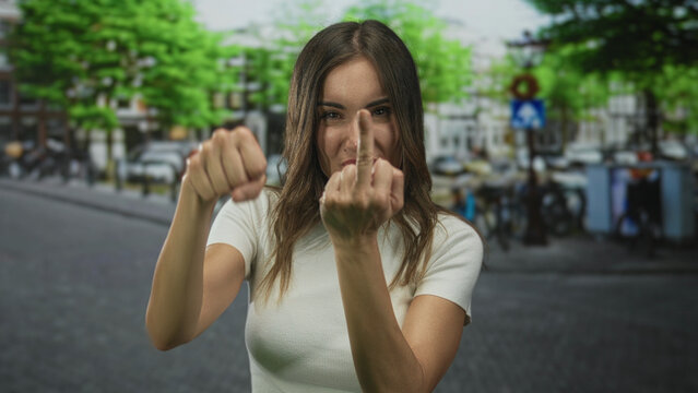 Woman flips middle finger on city street with blurred buildings under bright daylight; defiance.