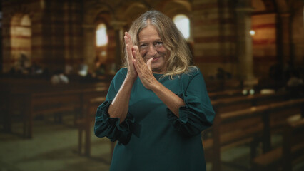 Fototapeta premium Senior woman claps hands inside a church among wooden pews, smiling with palms together and slight nod; gratitude reflection.