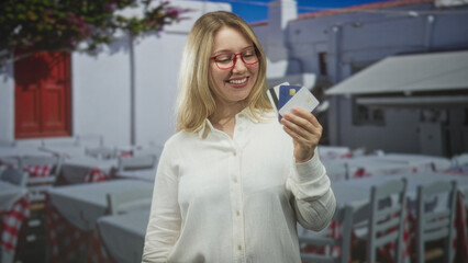 Fototapeta premium Woman holding creditcards in hand, smiling and presenting cards toward camera, blonde with red glasses, standing in a sunlit street cafe with tables and chairs; travel confidence.