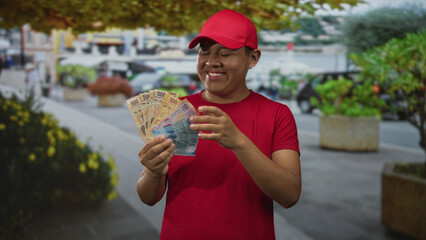 Fototapeta premium Hispanic delivery worker smiling while fanning mexican pesos on a busy street in red cap and shirt; happiness success.