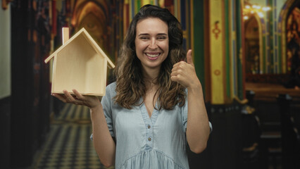 Fototapeta premium Woman holds wooden house model and gives thumbs up gesture in church building with colourful stained glass beyond; hope gratitude security.