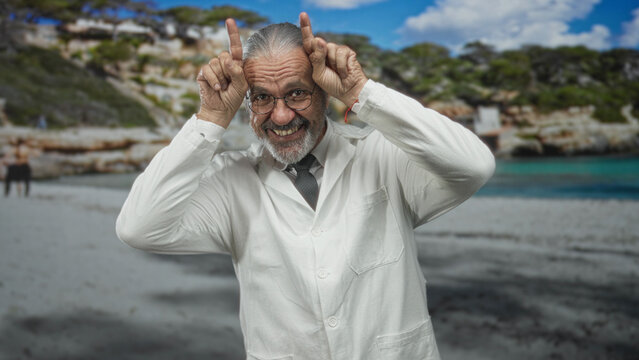 Scientist man with long gray hair and glasses in a white lab coat points index fingers up like horns while smiling on a sandy beach seashore; playful curiosity.