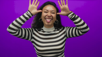 Young hispanic woman sticking tongue out with hands raised wearing hoop earrings and striped top in studio; playful mischief confidence.