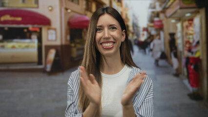 Fototapeta premium Young hispanic woman claps hands on busy city street with red awning and display windows outside shops; happiness.