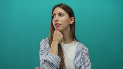 Fototapeta premium Young hispanic woman wearing striped shirt with hand on chin in teal studio scene; contemplation doubt.