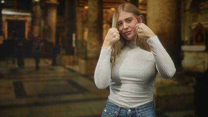 Fototapeta premium Woman clenches fists in a cathedral building, grimacing with fists raised near her face in jeans and sweater; defiance determination.