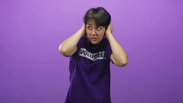 Woman volunteer in purple shirt covers ears with hands and grimaces in a studio while flinching from noise and looking distressed; anxiety overload.