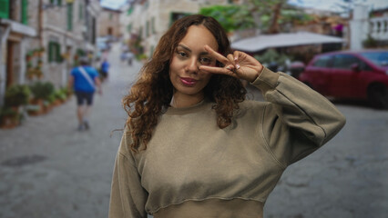 Fototapeta premium Woman flashing peace sign by eye on a cobblestone street with blurred passerby and parked car; youthful playful confidence.