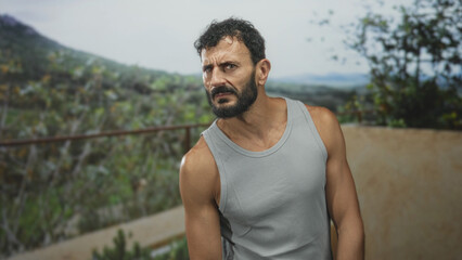 Fototapeta premium Man with beard and bare shoulder in a tank top tilts head and leans on a balcony railing on a building terrace overlooking hills; concern.