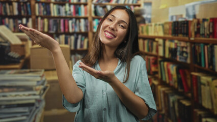 Hispanic young brunette woman smiling, palms up presenting gesture among bookshelves in a library building  curiosity. © Krakenimages.com