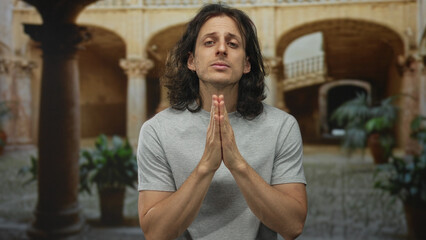 Man with long hair, hands clasped at chest, eyes closed in prayer before old town stone arches and...