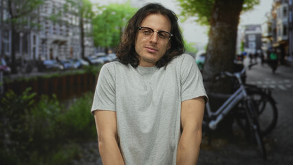 Fototapeta premium Young hispanic man with long hair and glasses shrugs shoulders showing bare forearms on a city street by a parked bicycle; shy introspection.