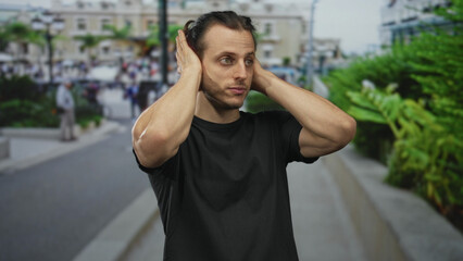 Fototapeta premium Man covers ears with hands on busy city street, bare forearms visible and mouth open in a grimace; frustration overload.