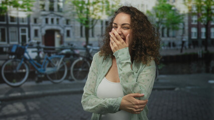 Fototapeta premium Woman laughing with hand covering mouth on amsterdam street by canal and bicycles; joy spontaneity now.