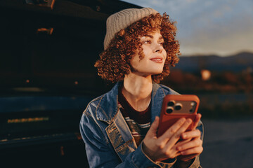 Curly woman wearing a beige hat and denim jacket holding smartphone while smiling outdoors during...