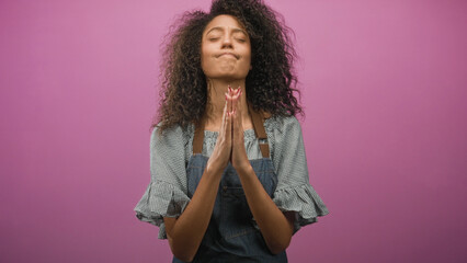 Obraz premium Woman with curly hair wearing denim apron, hands pressed together at chin in studio pink backdrop, closed eyes in praying gesture; hope.
