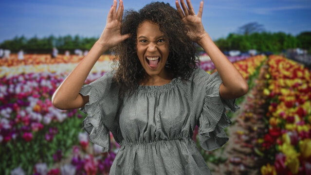 Teenage african american woman with curly hair crosses arms and pouts wearing gingham dress in studio with tulip backdrop; playful mischief.