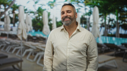 Naklejka premium Man standing with salt and pepper beard, hands relaxed at sides, neutral expression near pool loungers in building pool area; quiet contentment.