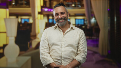 Obraz premium Man with beard smiling, hands clasped while seated in a hotel building lobby with lamp and curtains; contentment.