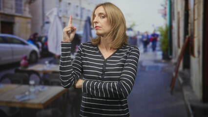 Fototapeta premium Woman points finger upward with folded arms at street cafe table, looking to the side; impatience urgency.