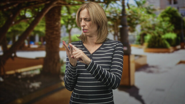 Woman inspects a fingernail on her hand in a busy street plaza, using the other hand to point and closely examine a possible hangnail or small injury; disgust concern.