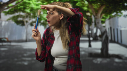 Woman painter in red plaid shirt holds paintbrush upright and shields eyes with hand on forehead while peering down a sunlit street  curiosity discovery. © Krakenimages.com