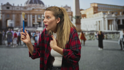 Fototapeta premium Woman holding paintbrush, points thumb to chest in front of building and basilica on crowded plaza; surprise curiosity.