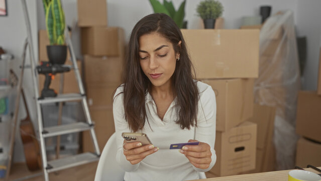 Woman holding smartphone and creditcard with hands visible while sitting among moving boxes in a building; budget planning focused.