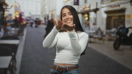 Fototapeta premium Young hispanic woman pinching fingers and smiling with visible hands and casual white top on a bustling pedestrian street lined with cafes and scooters; playful joy.