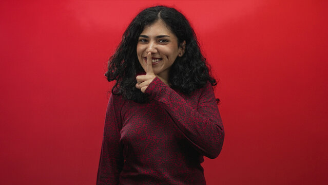 Young arab brunette woman finger to lips for silence in red studio with curly hair and dimpled smile; playful mischief.