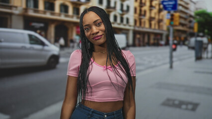 Fototapeta premium Black woman smiling at camera, showing midriff and long braids, wearing pink crop top while standing on a city street with passing van; confident casual.