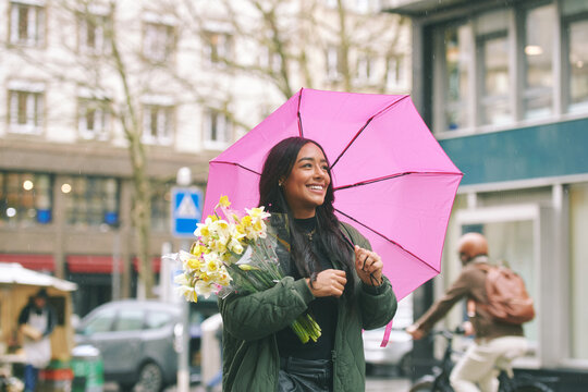 Happy young Latina woman holding flowers under pink umbrella on rainy city street, stylish female enjoying spring bouquet while walking in urban European setting