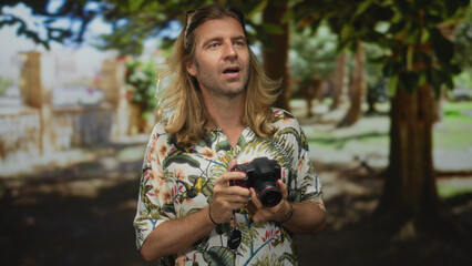 Man wearing tropical shirt and sunglasses holds camera to eye while framing a shot beneath leafy...