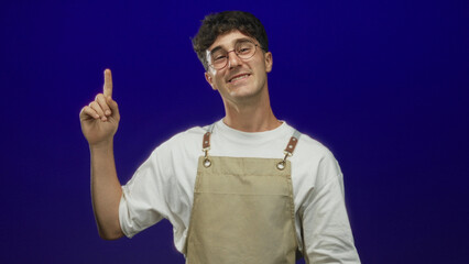 Naklejka premium Man pointing index finger in blue studio, smiling and wearing beige apron and round glasses; playful curiosity.