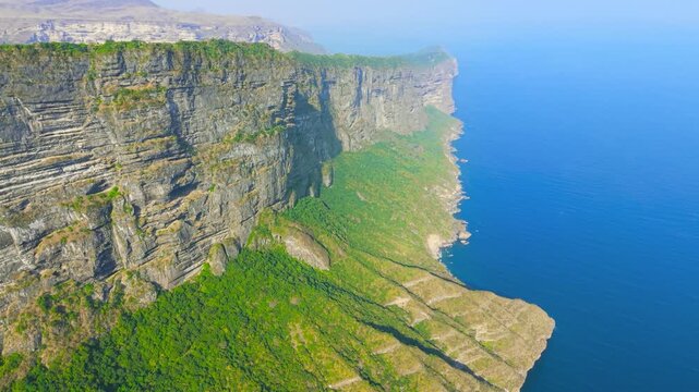 Aerial view of high rocky cliffs covered in green vegetation overlooking the blue sea at Jabal Shaat on the border of Oman and Yemen.