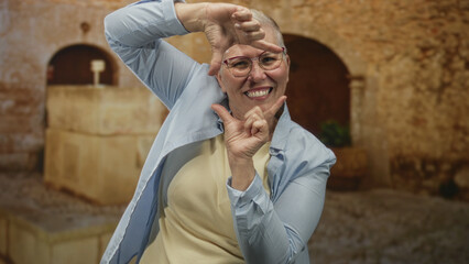 Obraz premium Woman with short grey hair and glasses frames face with hands on street in old town, smiling broadly in a light shirt; joyful confidence.