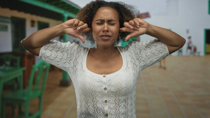 Woman pressing fingers into ears and grimacing standing in a sunlit colonial building courtyard;...