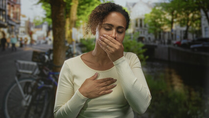 Fototapeta premium Woman covering mouth with hand and clutching chest on street by canal and parked bicycle, heartattack; distress anxiety worry.