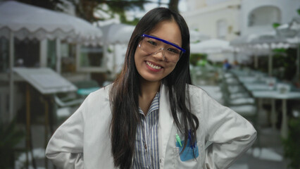 Fototapeta premium Woman smiling in a scientist uniform with safety glasses points at the camera while enjoying an outdoor terrace at a restaurant, blending science and leisure.