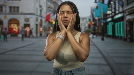 Fototapeta premium Young african american woman pressing hands to cheeks with closed eyes on a busy city street lined with shops; anxiety introspection.