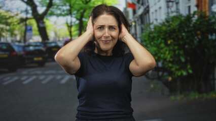 Fototapeta premium Woman covers ears with hands on a street, eyes shut and grimacing amid traffic and parked cars; distress frustration.